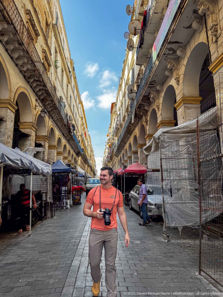 Visitor with camera standing in a broad market street lined by repeating arcades, balconies, and covered stalls.