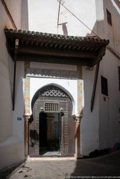 Arched palace entrance beneath a timber canopy, with columns, carved doorway, and colorful tile panels set in white walls.