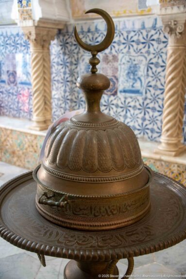 Ornate circular metal object with lobed domed cover, engraved bands, and crescent finial, displayed before blue patterned tiles.