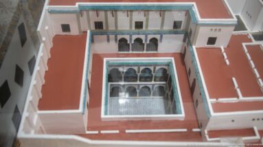 Overhead architectural model with a square central courtyard, arcaded galleries, white walls, red roofs, and turquoise trim.