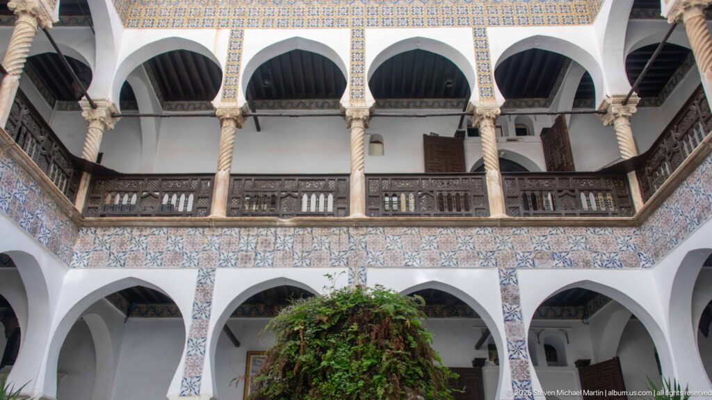 Upward courtyard view of two-story white arcades with spiral columns, patterned tile bands, and dark wooden upper-gallery railings.