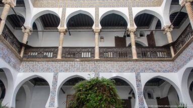 Upward courtyard view of two-story white arcades with spiral columns, patterned tile bands, and dark wooden upper-gallery railings.