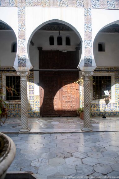 Large carved wooden double door beneath white arches, framed by spiral columns and colorful patterned tilework in the courtyard.