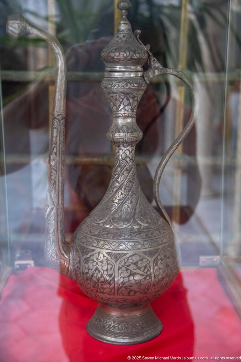 Ornate engraved metal ewer with bulbous body, long spout, curved handle, and finial displayed behind glass on red fabric.