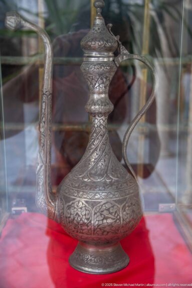 Ornate engraved metal ewer with bulbous body, long spout, curved handle, and finial displayed behind glass on red fabric.