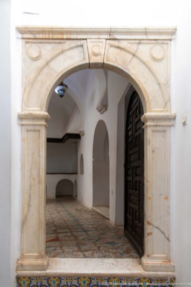 Pale stone rounded portal opening into a white vaulted room with tiled floor, wall arches, and a dark wooden door.