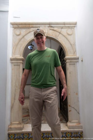 Man in a green shirt and cap standing beneath a pale stone arched doorway inside Dar Mustapha Pacha.