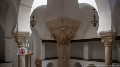 Large pale stone column with carved capital supporting intersecting white arches, with hanging lamps in the background.