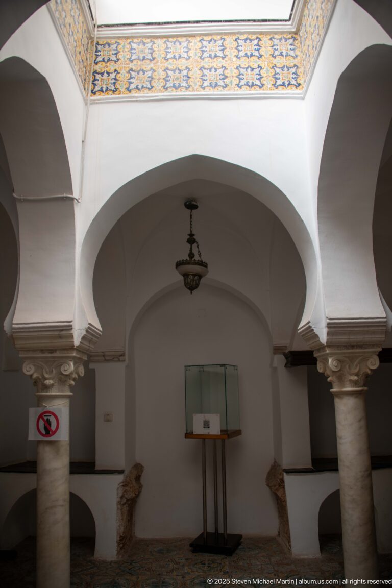 White interior arches on pale stone columns, with a hanging lamp, wall display case, and small exhibit object below.