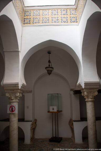 White interior arches on pale stone columns, with a hanging lamp, wall display case, and small exhibit object below.