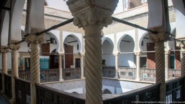 Upper-gallery view through spiral columns and dark railing toward tiled two-story courtyard arcades and wooden doors.
