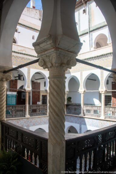 Close upper-level view of a spiral-fluted stone column and carved capital, with white courtyard arches and tile bands beyond.