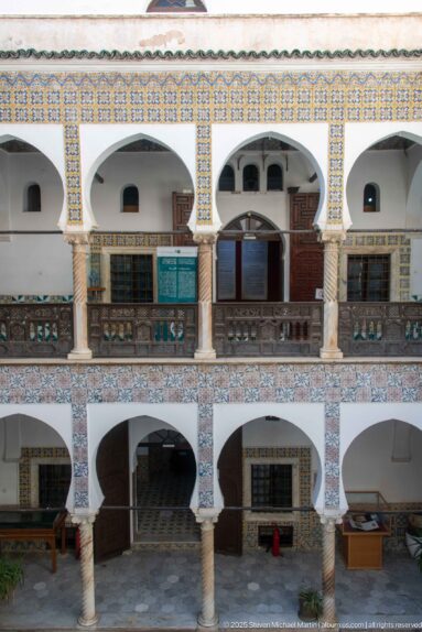 Frontal view of a two-story courtyard wall with white arches, slender columns, dark wooden railings and doors, and patterned tile bands.