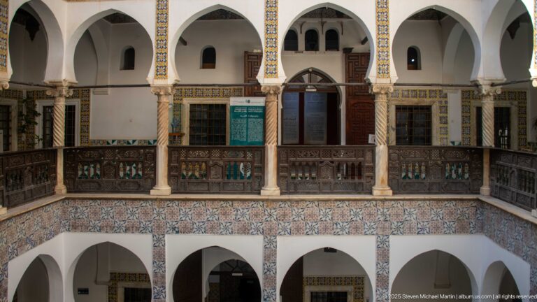Upper courtyard gallery with white arches, columns, dark wooden balustrade and doors, above a broad band of patterned tiles.