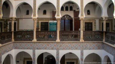 Upper courtyard gallery with white arches, columns, dark wooden balustrade and doors, above a broad band of patterned tiles.