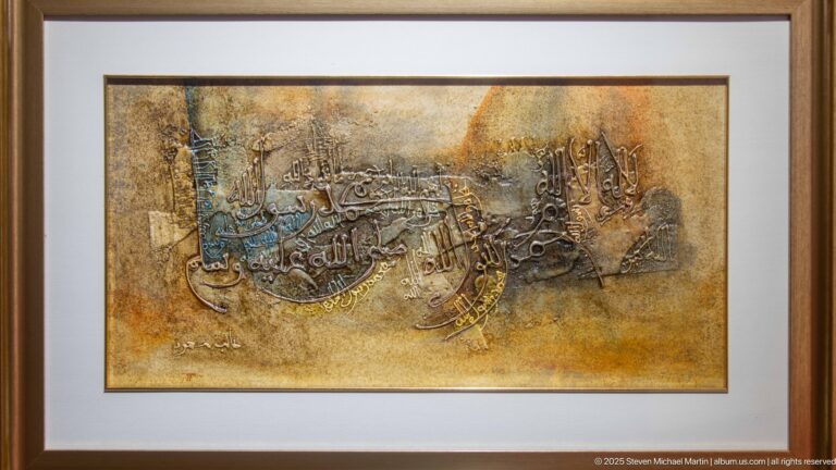 Framed Arabic calligraphic artwork with layered dark script across a textured gold-brown background.