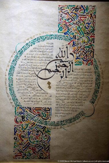 Arabic calligraphic artwork with circular fine text, bold black central script, turquoise lettering, and multicolored illuminated ornament.