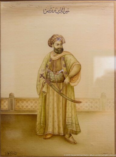 Portrait of a turbaned man in long robes with a curved sword, standing before a faint architectural background with Arabic inscriptions.