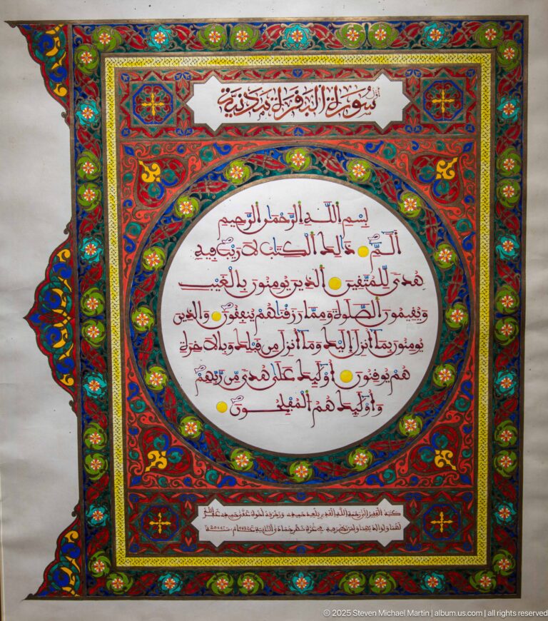Arabic manuscript-style page with red text in a white oval, surrounded by elaborate red, blue, green, and gold illuminated borders.