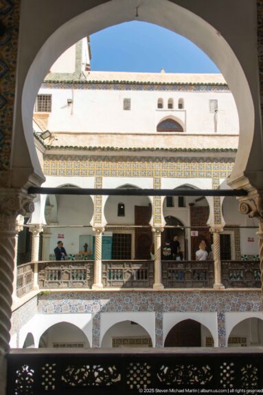Courtyard seen through a broad white arch, with opposite tiled galleries, dark railings, visitors, and blue sky beyond.