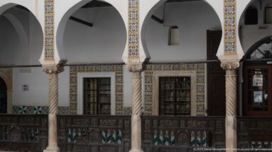 Upper gallery with two spiral stone columns, barred windows framed by blue-and-yellow tiles, and a dark carved wooden railing.