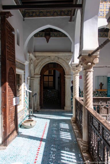 Sunlit palace gallery with spiral columns, carved wooden railing, patterned floor, and a large arched doorway at the far end.