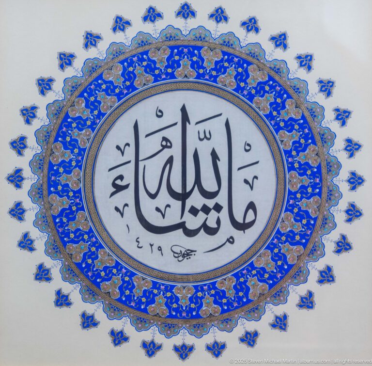 Large black Arabic calligraphy in a pale oval surrounded by dense cobalt-blue and gold floral illumination and small outer motifs.