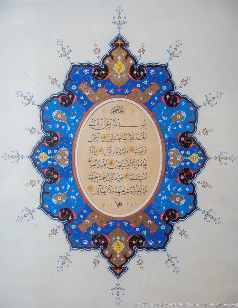 Circular Arabic text surrounded by a blue, red, gold, and turquoise lobed illuminated medallion on a pale background.