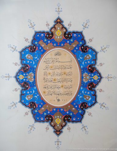 Circular Arabic text surrounded by a blue, red, gold, and turquoise lobed illuminated medallion on a pale background.