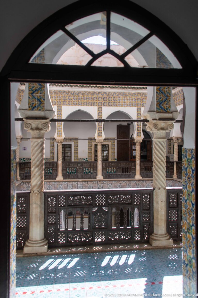 Shaded upper gallery framed by a dark arch, with spiral columns, carved wooden railing, tiled courtyard walls, and patterned sunlight.