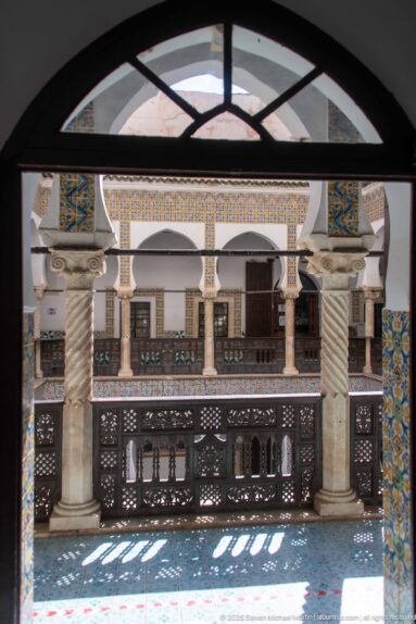 Shaded upper gallery framed by a dark arch, with spiral columns, carved wooden railing, tiled courtyard walls, and patterned sunlight.
