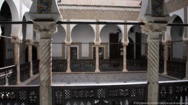 Spiral-fluted columns and dark carved railings frame a view across a tiled, arcaded interior courtyard gallery.