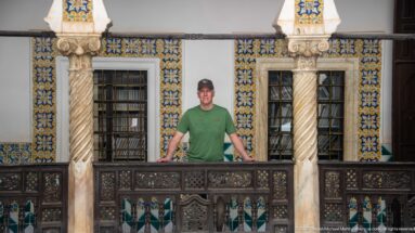 Man in green shirt and cap standing behind a carved wooden gallery railing between spiral columns and tile-framed windows.