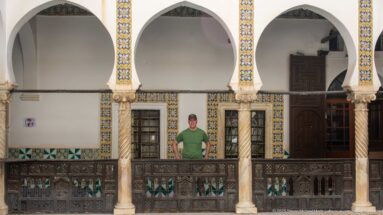 Wide upper-gallery elevation with a visitor behind carved railing, three white arches, spiral columns, tiled window borders, and dark door.