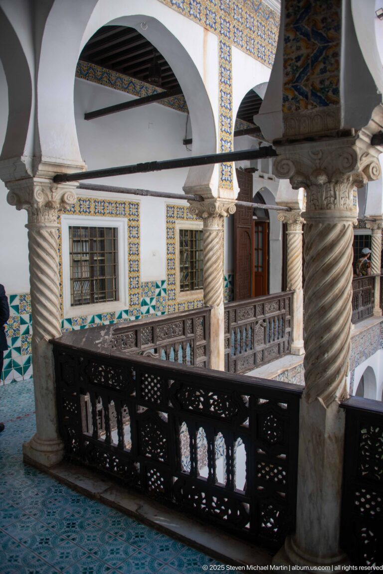 Oblique upper gallery with dark carved railing, spiral columns, white arches, tiled window borders, and a wooden door.