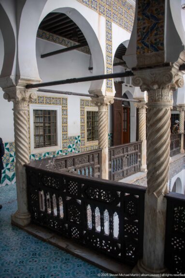 Oblique upper gallery with dark carved railing, spiral columns, white arches, tiled window borders, and a wooden door.
