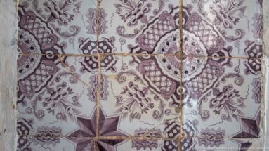 Close view of pale ceramic tiles with purple floral medallions, leafy ornament, pointed stars, cracks, and worn grout lines.