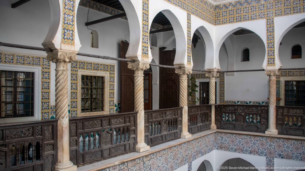 Corner view of upper courtyard arcades with white arches, spiral columns, patterned tile borders, windows, doors, and dark wooden railings.
