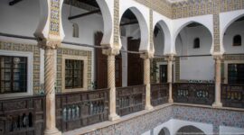 Corner view of upper courtyard arcades with white arches, spiral columns, patterned tile borders, windows, doors, and dark wooden railings.