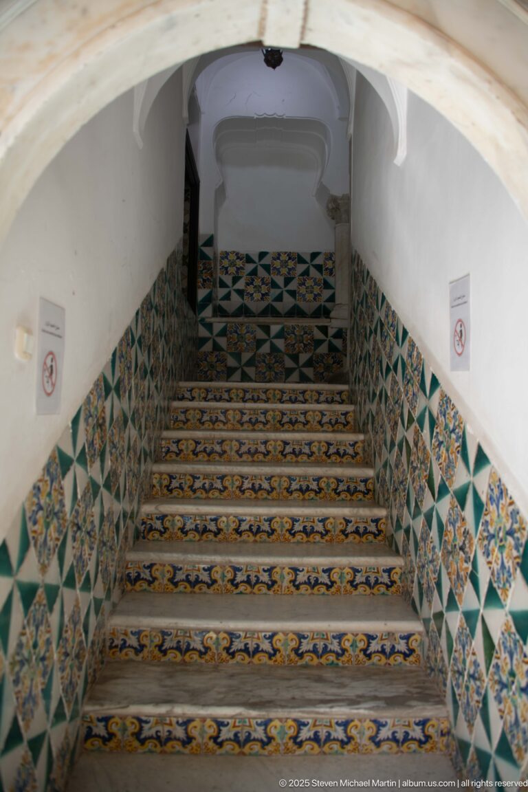 Interior staircase beneath a white vault, with patterned tile risers and green, blue, yellow, and white geometric wall tiles.