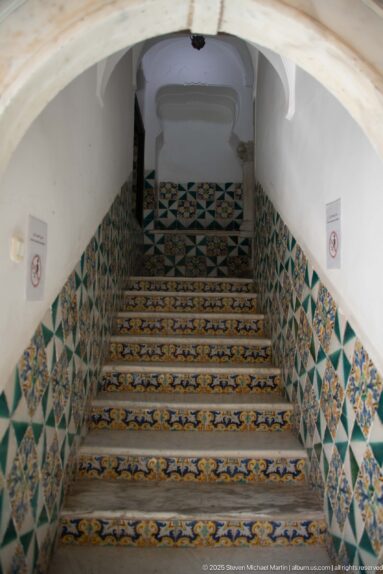 Interior staircase beneath a white vault, with patterned tile risers and green, blue, yellow, and white geometric wall tiles.