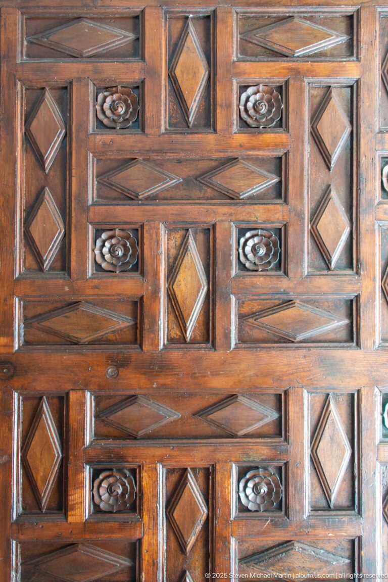 Close view of dark carved woodwork with raised rectangles, diamond insets, diagonal bands, and small rosette motifs.