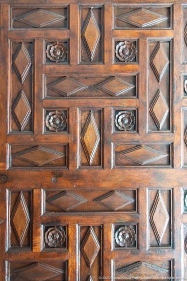 Close view of dark carved woodwork with raised rectangles, diamond insets, diagonal bands, and small rosette motifs.