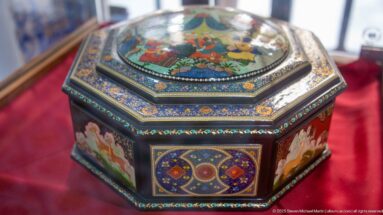 Octagonal dark decorative box with miniature painted panels, intricate multicolored borders, and a figurative scene on the raised lid.