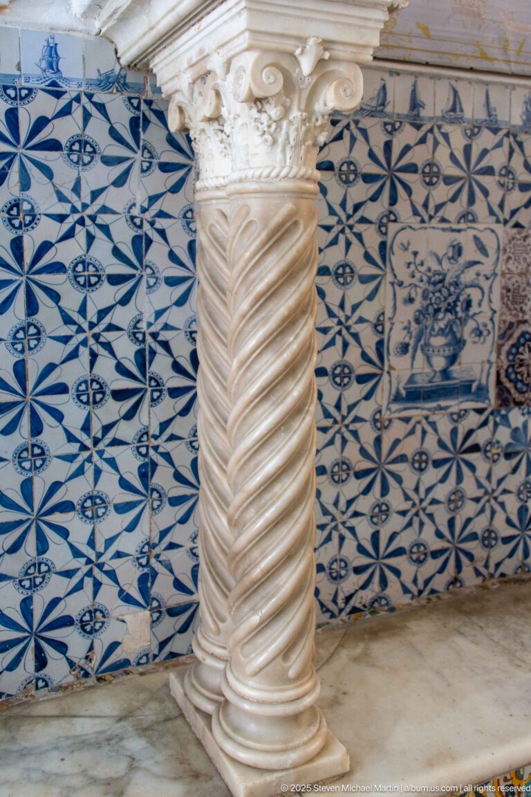 Spiral-fluted pale stone column with carved capital standing before blue-and-white patterned ceramic wall tiles.