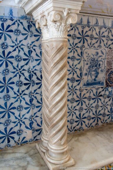 Spiral-fluted pale stone column with carved capital standing before blue-and-white patterned ceramic wall tiles.