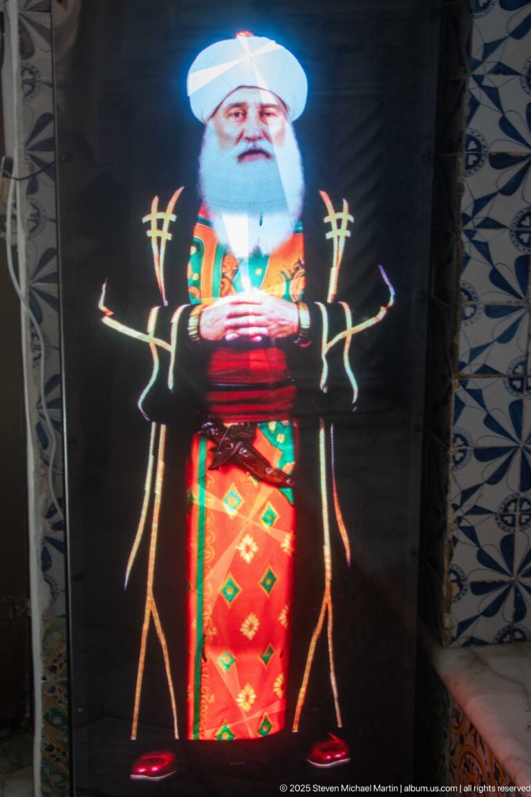 Glowing digital figure of a white-bearded man in a white turban and colorful patterned robes against a dark display.