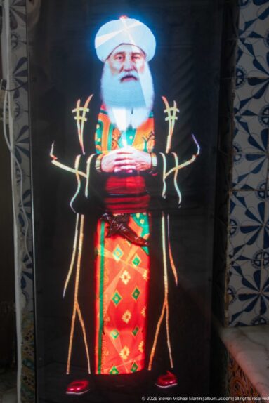Glowing digital figure of a white-bearded man in a white turban and colorful patterned robes against a dark display.