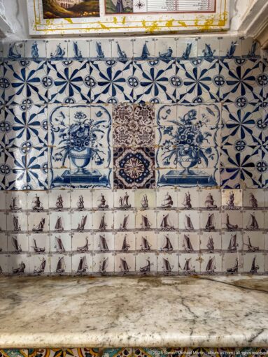 Wall above a marble ledge with blue-and-white floral vase tiles, central ornament, and rows of small sailing-ship tiles.