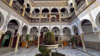 Wide courtyard with central fountain and plant, surrounded by two levels of white arches, spiral columns, tiles, and dark wooden galleries.